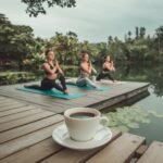 Yoga Session Taking Place. A photo of a yoga session taking place in a serene outdoor setting. There are three pretty models performing yoga poses. In the foreground, a cup of black coffee is placed on a wooden platform. The background contains lush greenery and a calm lake. The sky is overcast.